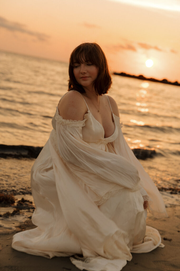 Woman kneeling in the sand wearing a flowing white dress during a sunset portrait session