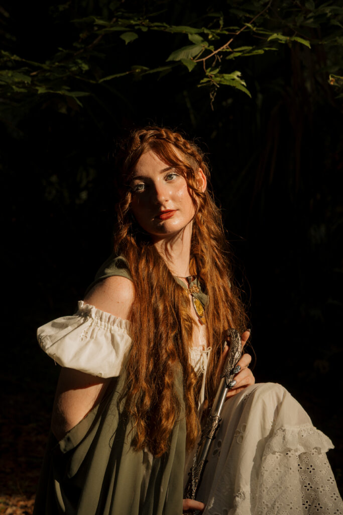 Woman seated in the woods wearing a white dress and green cape during a medieval inspired portrait session