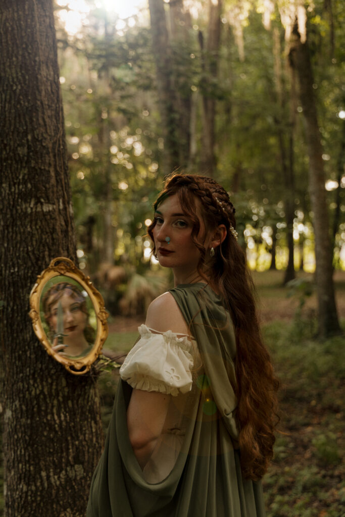 Woman leaning from behind a tree holding an ornate mirror during a creative fantasy portrait session