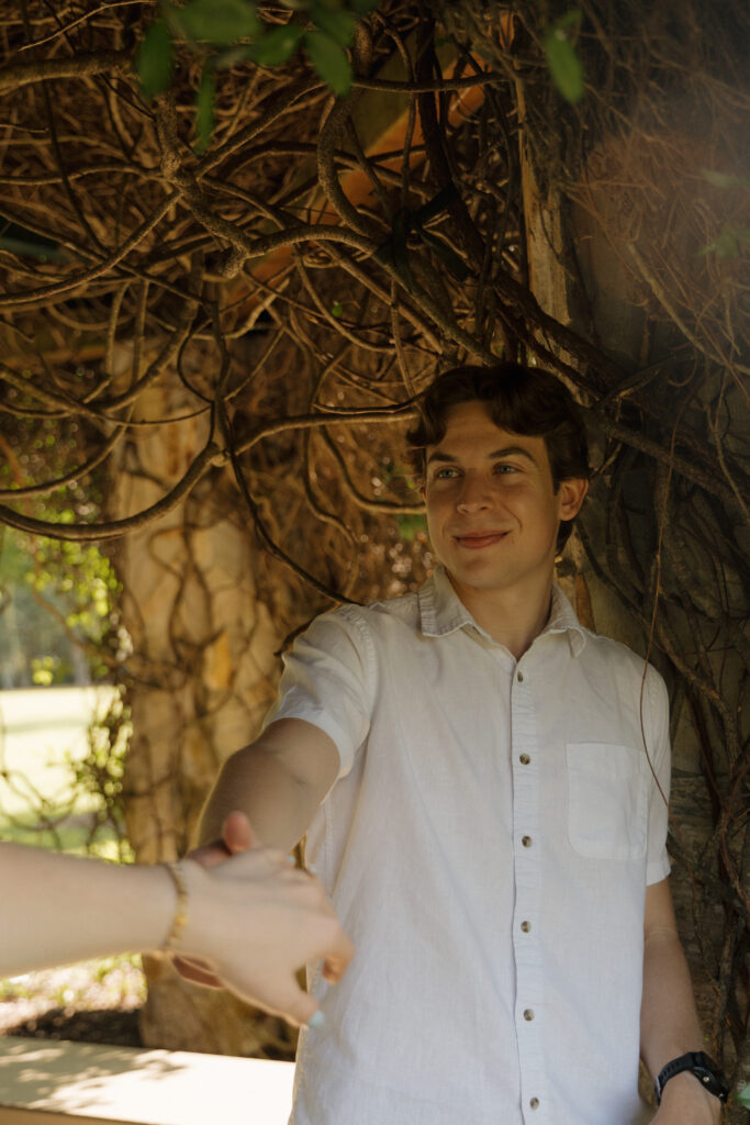 Groom to be reaching for his fiancé’s hand beneath hanging vines at Sholom Park