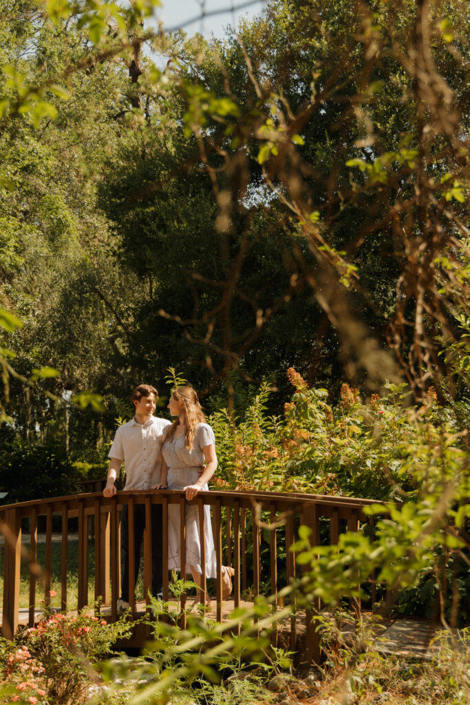 Engaged couple standing together on a wooden bridge surrounded by gardens at Sholom Park